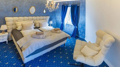 Elegantes Doppelzimmer in Blau mit edlem Kronleuchter, gepolstertem Bett und stilvollen Vorhängen.